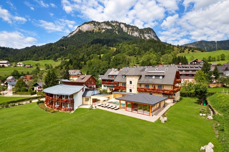 Hotel Hechl in Tauplitz mit Sonnenterrasse und grüner Wiese vor bewaldeter Bergkulisse.