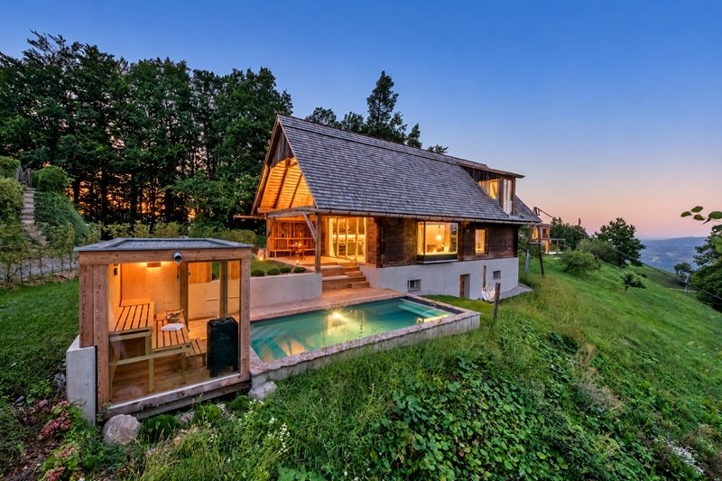 PURESLeben-Ferienhaus in der Südsteiermark mit beleuchtetem Außenpool und verglaster Sauna in der Abenddämmerung.